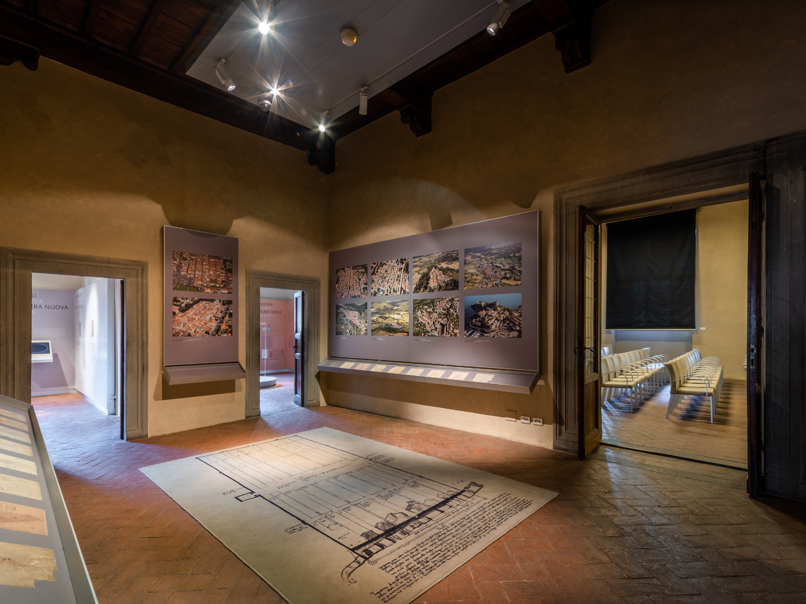 Ein Raum im Museo delle Terre Nuove. Einige Zeichnungen und Luftaufnahmen sind zu sehen. Rechts sieht man den Eingang zu einem Raum mit einigen Stühlen.